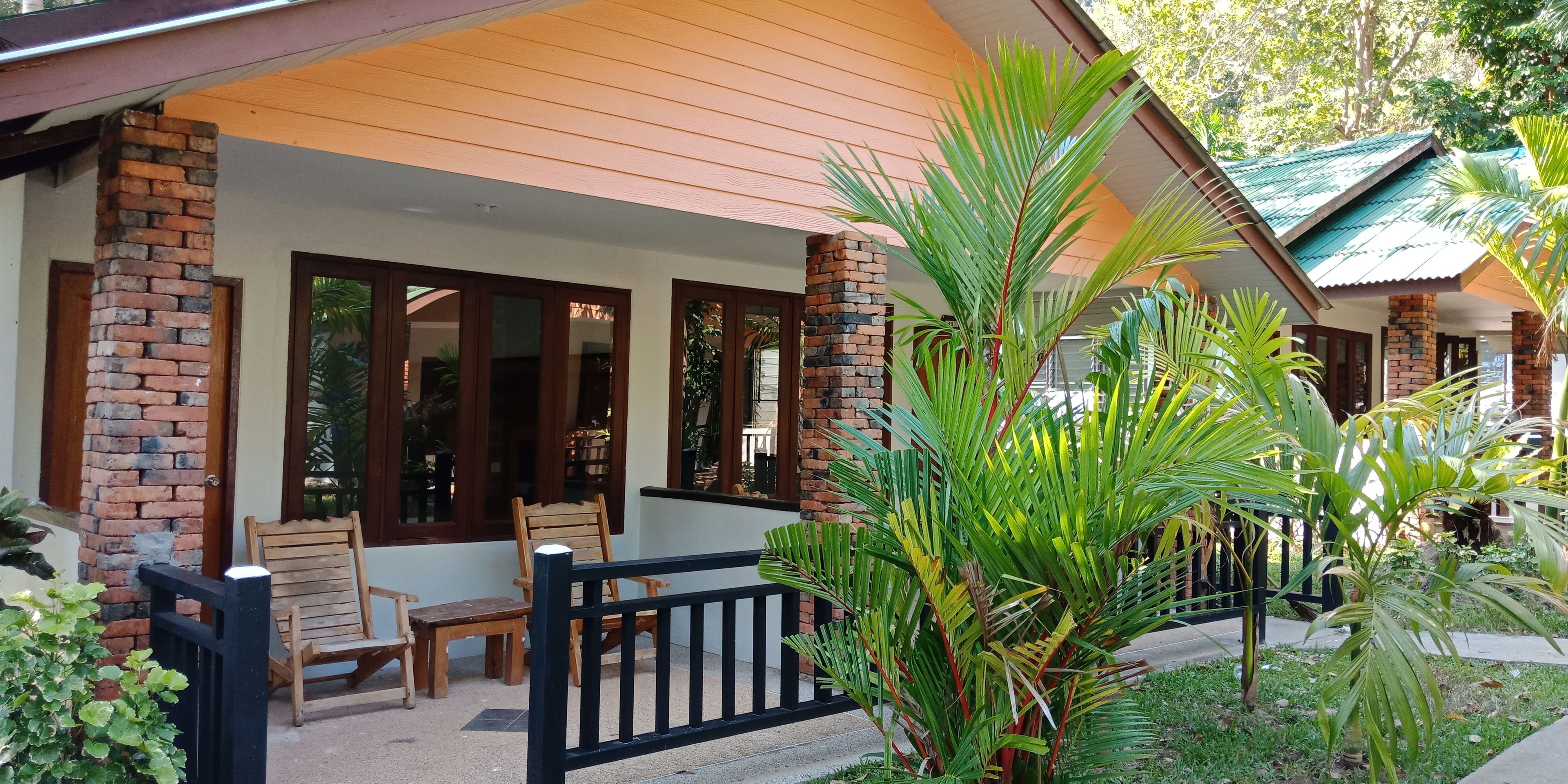 A covered outdoor seating area with wooden chairs and a black fence, surrounded by lush greenery and a brick pillar.