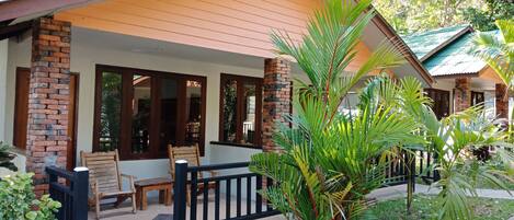 A covered outdoor seating area with wooden chairs and a black fence, surrounded by lush greenery and a brick pillar.