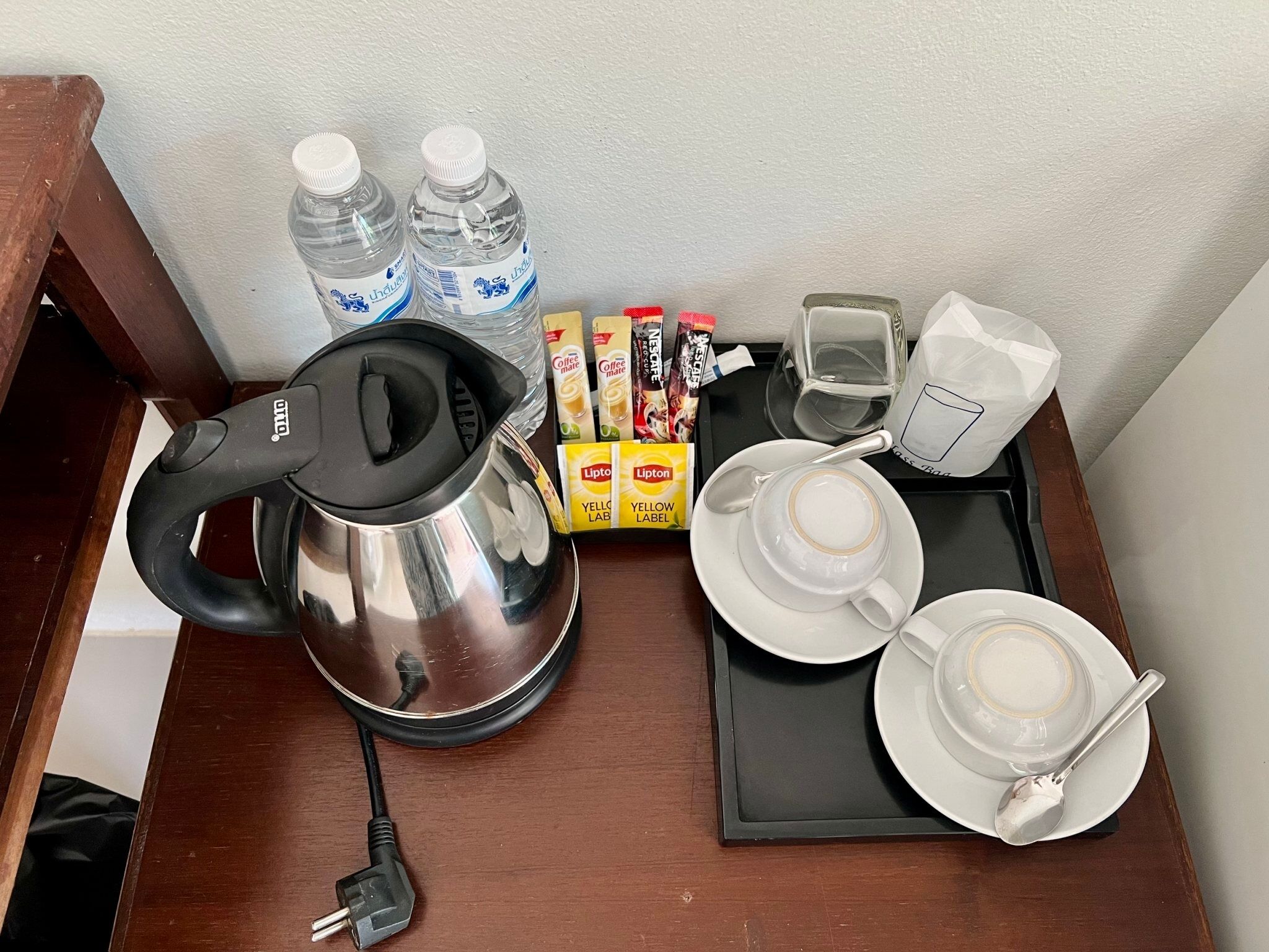 Una bandeja con una tetera, bolsitas de té Lipton, té Lipton, sobres de crema para café, un vaso y una taza sobre una mesa de madera.