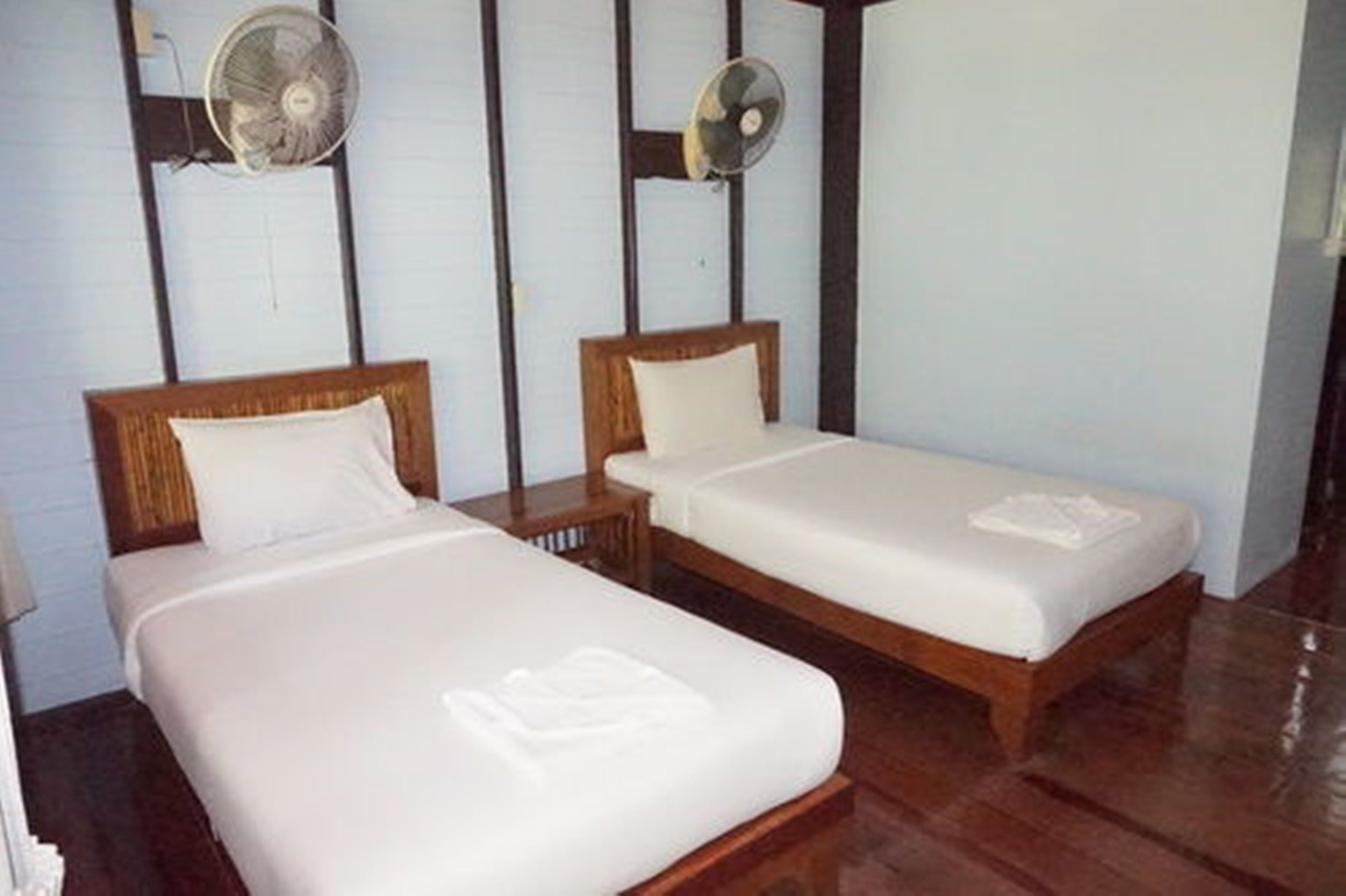 Two single beds with wooden headboards and white bedding, a wooden floor, and two wall-mounted fans.