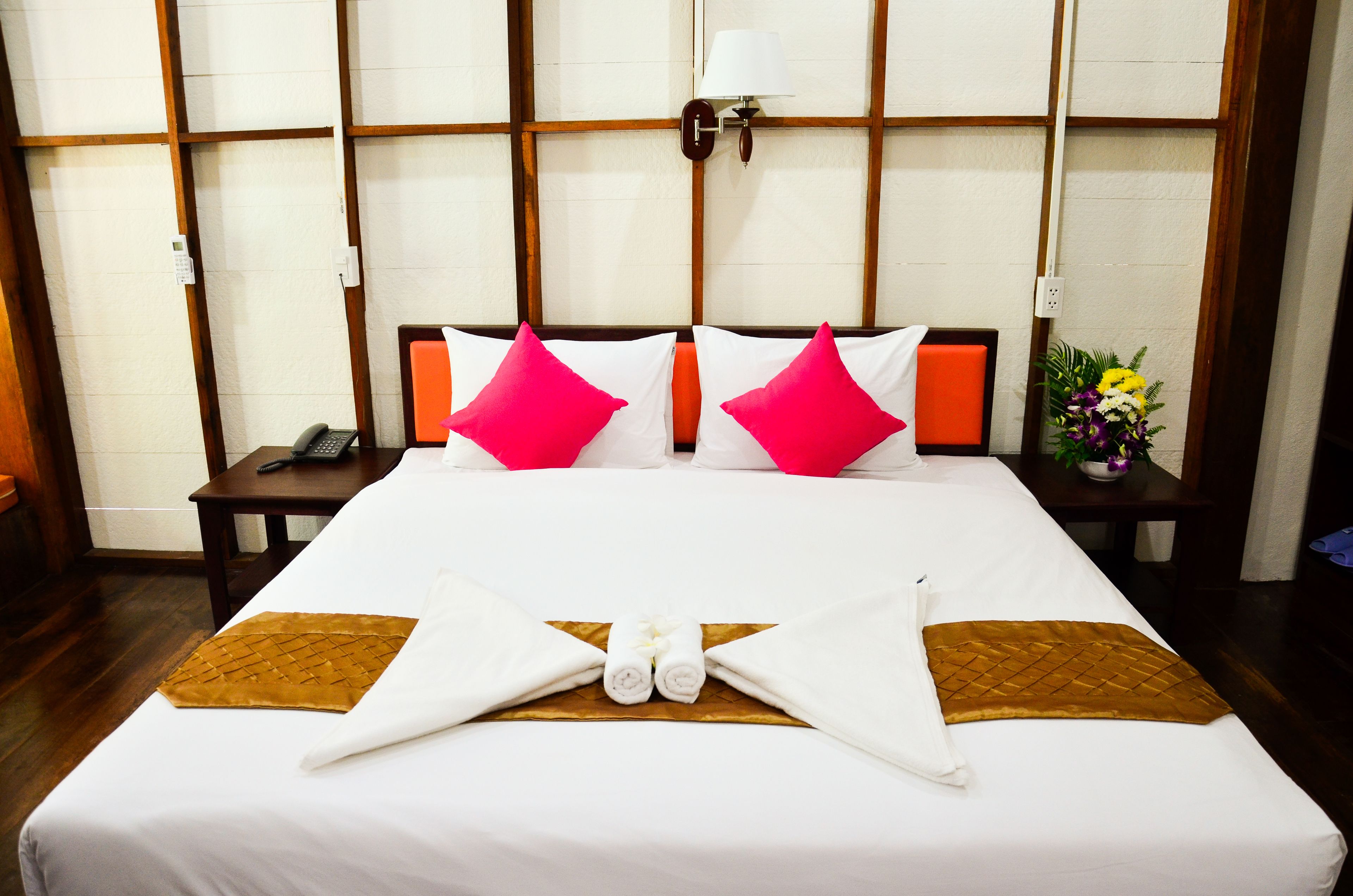 Una cama bien hecha con almohadas blancas y rosadas, una almohada decorativa marrón y una disposición de toallas en el centro.