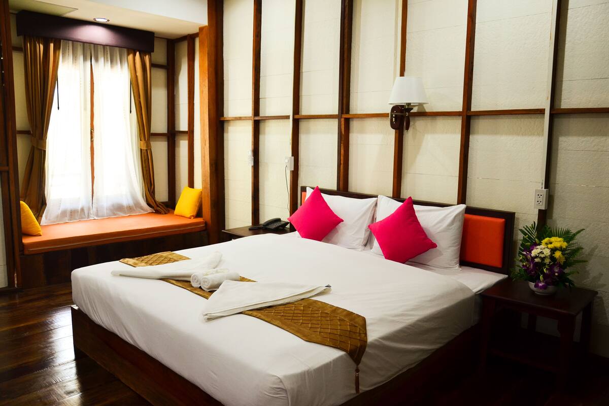 Una habitación de hotel con una cama grande, una mesita de noche con lámpara, una ventana con cortinas y una pequeña mesa con un jarrón de flores.