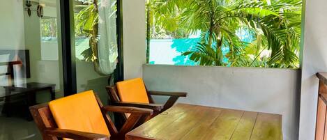 Une table en bois et deux chaises dotées de coussins orange, dans une pièce offrant une vue sur un feuillage verdoyant luxuriant, visible à travers une fenêtre.