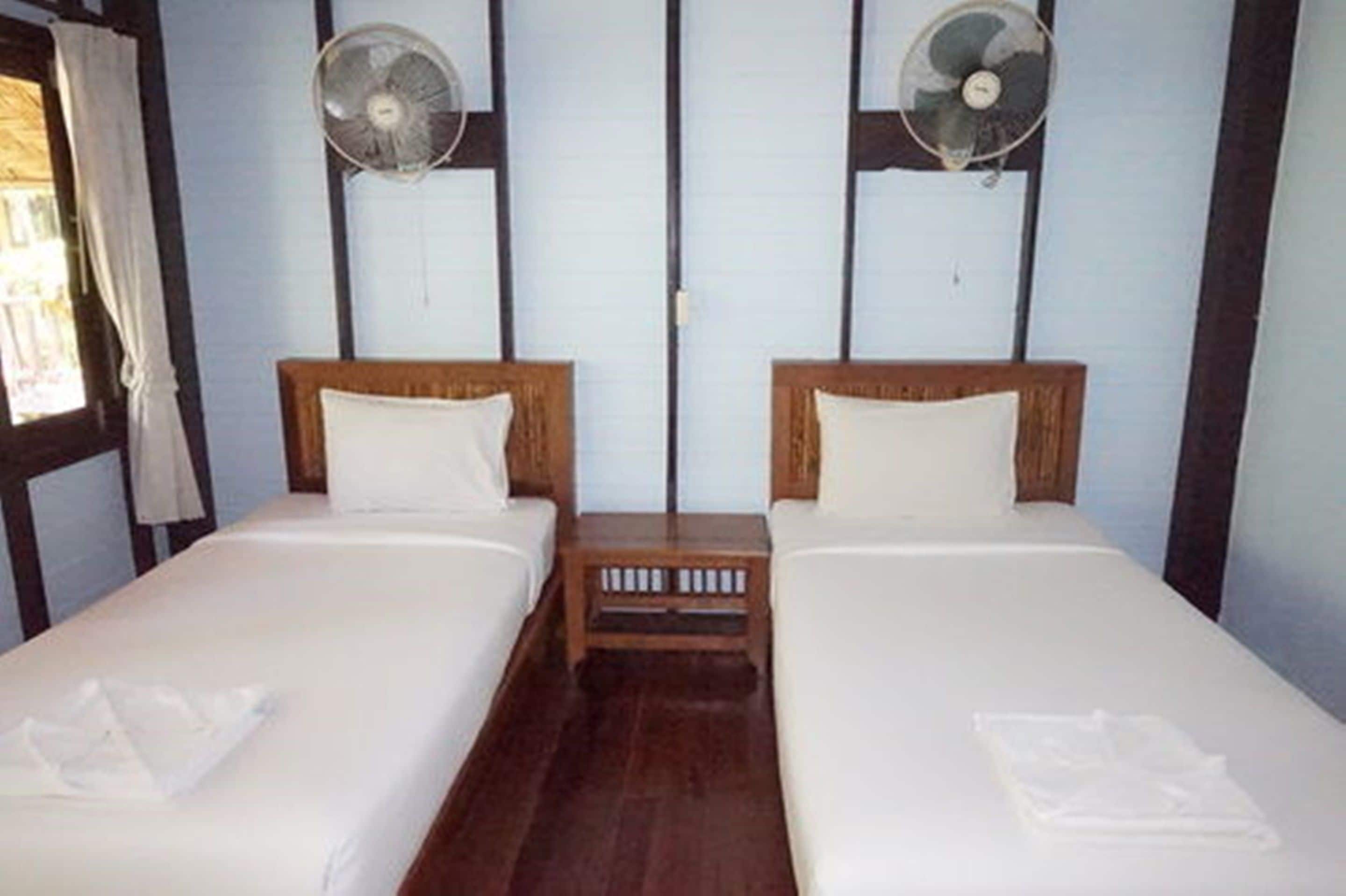 Une chambre avec deux lits simples, des têtes de lit en bois, du linge de lit blanc et deux ventilateurs fixés au mur.