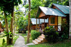A bungalow-style hotel with a wooden exterior and a green roof, surrounded by lush greenery and palm trees.
