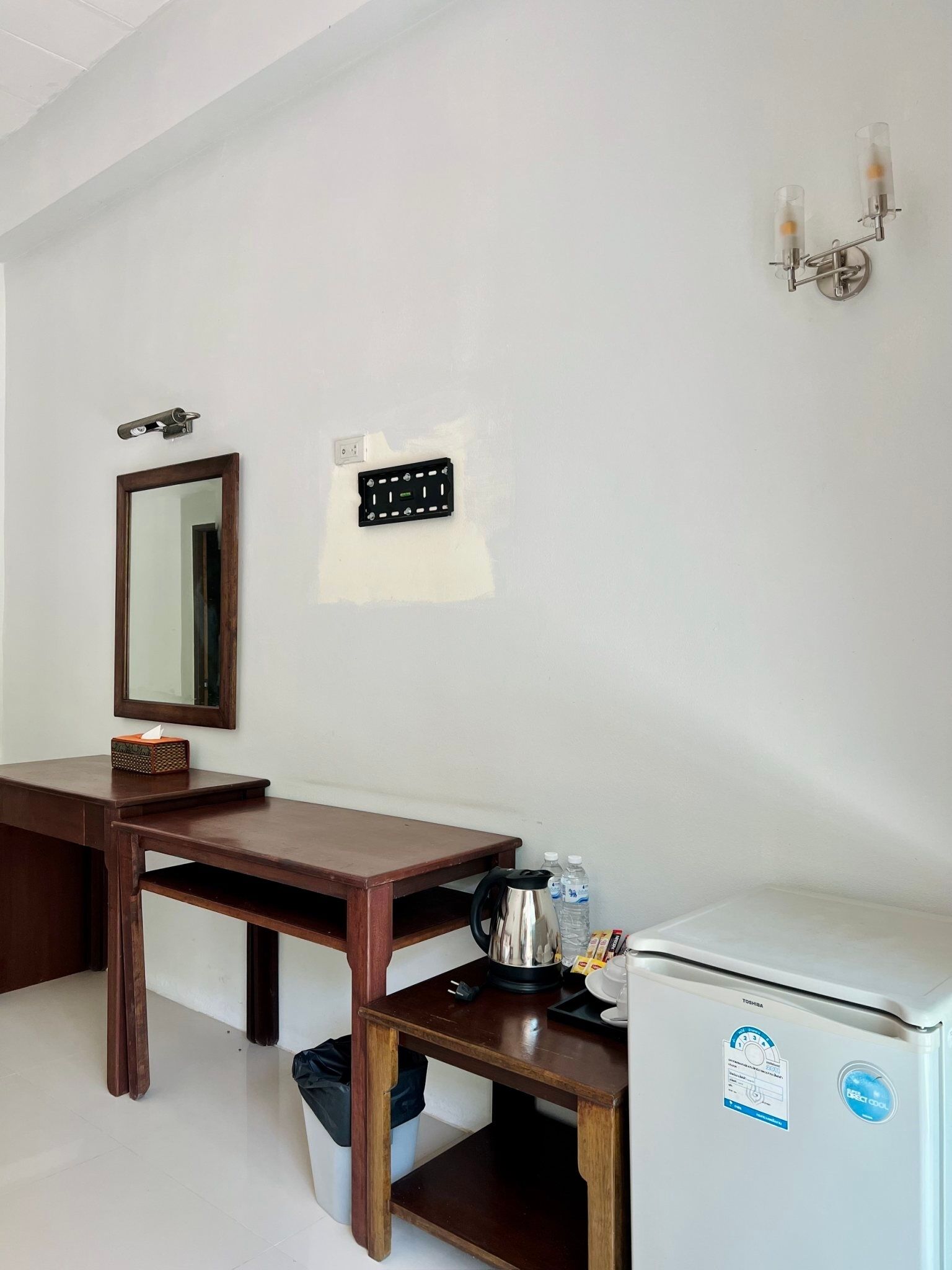 Habitación de hotel con un escritorio de madera, un espejo, una lámpara de pared, un termostato y un refrigerador.