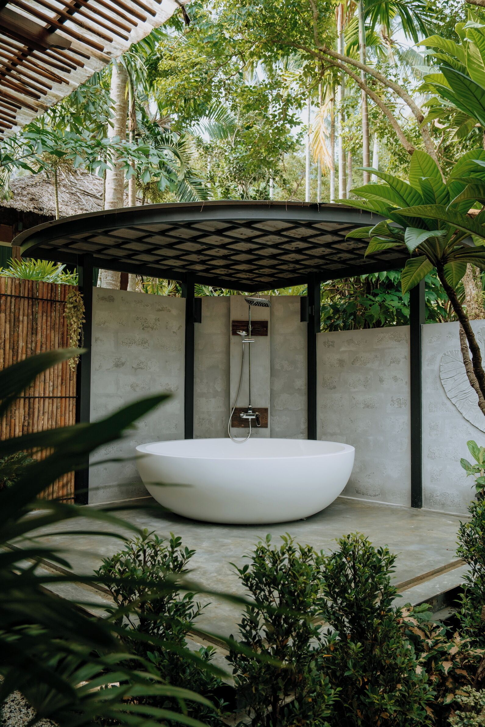 A modern outdoor bathroom with a large white bathtub, a shower, and a concrete wall.
