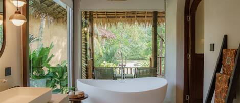 A modern bathroom with a freestanding bathtub, a large window, and a view of a tropical garden.