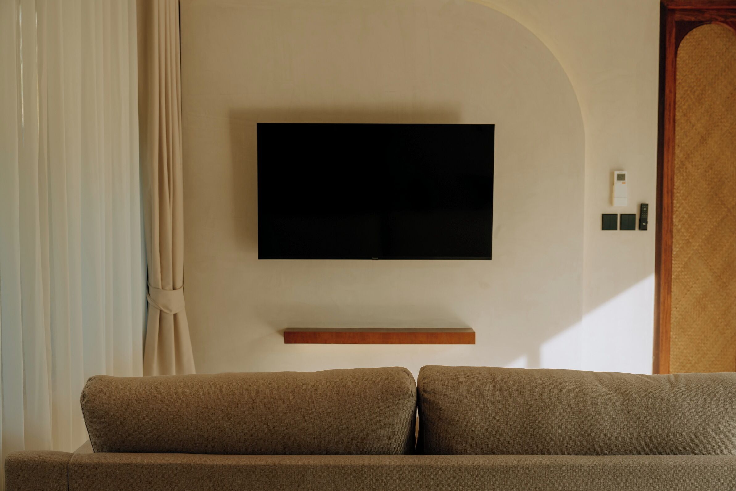 Un salon avec un téléviseur à écran plat fixé au mur, une étagère en bois en dessous et un canapé beige.