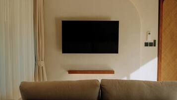 Un salon avec un téléviseur à écran plat fixé au mur, une étagère en bois en dessous et un canapé beige.