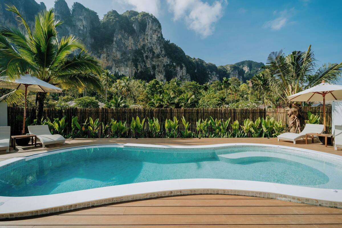 Una piscina con sillones y sombrillas, rodeada de exuberante vegetación y una valla de bambú, con montañas al fondo.