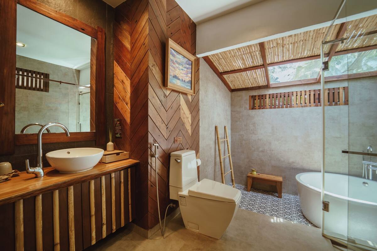 Un baño moderno con pared de madera en patrón de espiga, un espejo grande, un lavabo sobre encimera, un inodoro, un banco y una bañera con mampara de vidrio.
