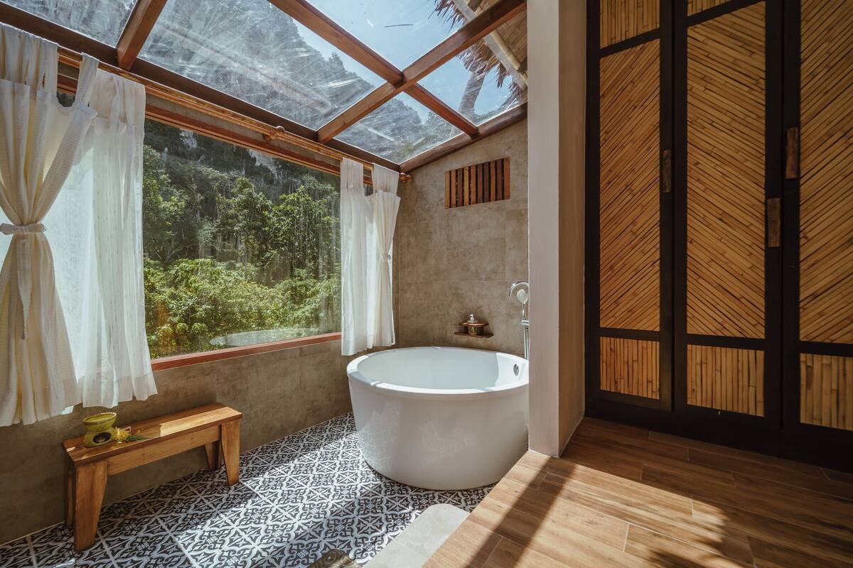 Un baño moderno con una bañera independiente, un banco de madera, baldosas de suelo estampadas y un amplio ventanal con cortinas.