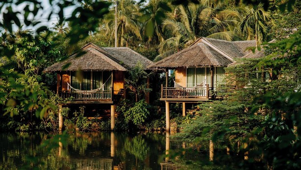 Two thatched-roof overwater bungalows with wooden decks, surrounded by lush greenery and palm trees.