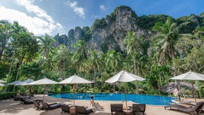 A poolside area with lounge chairs, umbrellas, and a view of a cliff and palm trees.
