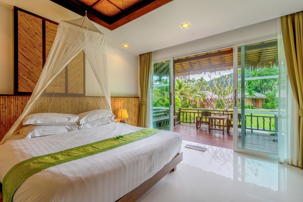 A bedroom with a bed, a canopy, a balcony with a table and chairs, and a view of greenery.