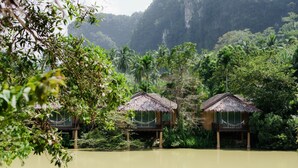 A row of thatched-roof huts by a calm lake, surrounded by dense tropical foliage and a steep cliff in the background.
