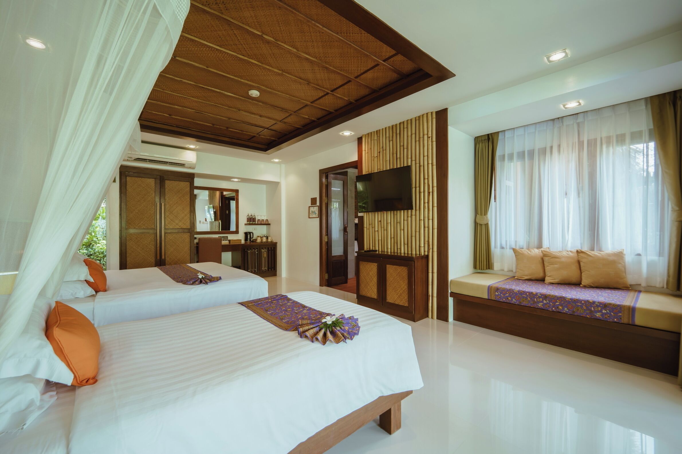 Quarto de hotel com duas camas, televisão fixada na parede, área de estar com almofadas e uma janela grande com cortinas.