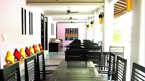 Un amplio comedor con mesas y sillas largas, ventiladores de techo y vista a la habitación contigua.