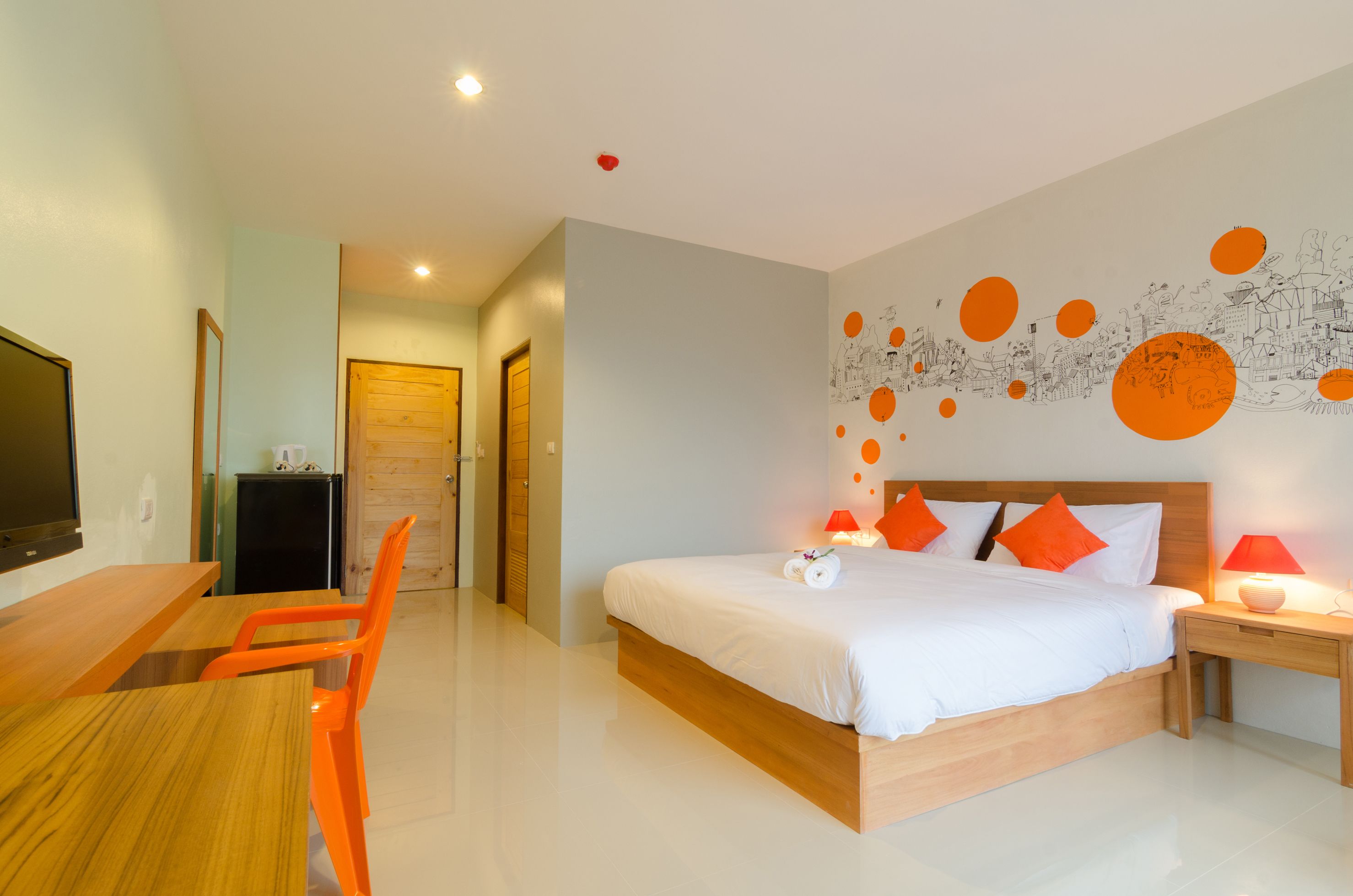 A modern hotel room with a wooden bed, orange chairs, a desk, and a TV.