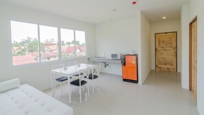 A modern, bright room with a dining area, a small kitchen, and a large window offering a view of buildings outside. - Aonang Miti Resort (Krabi)