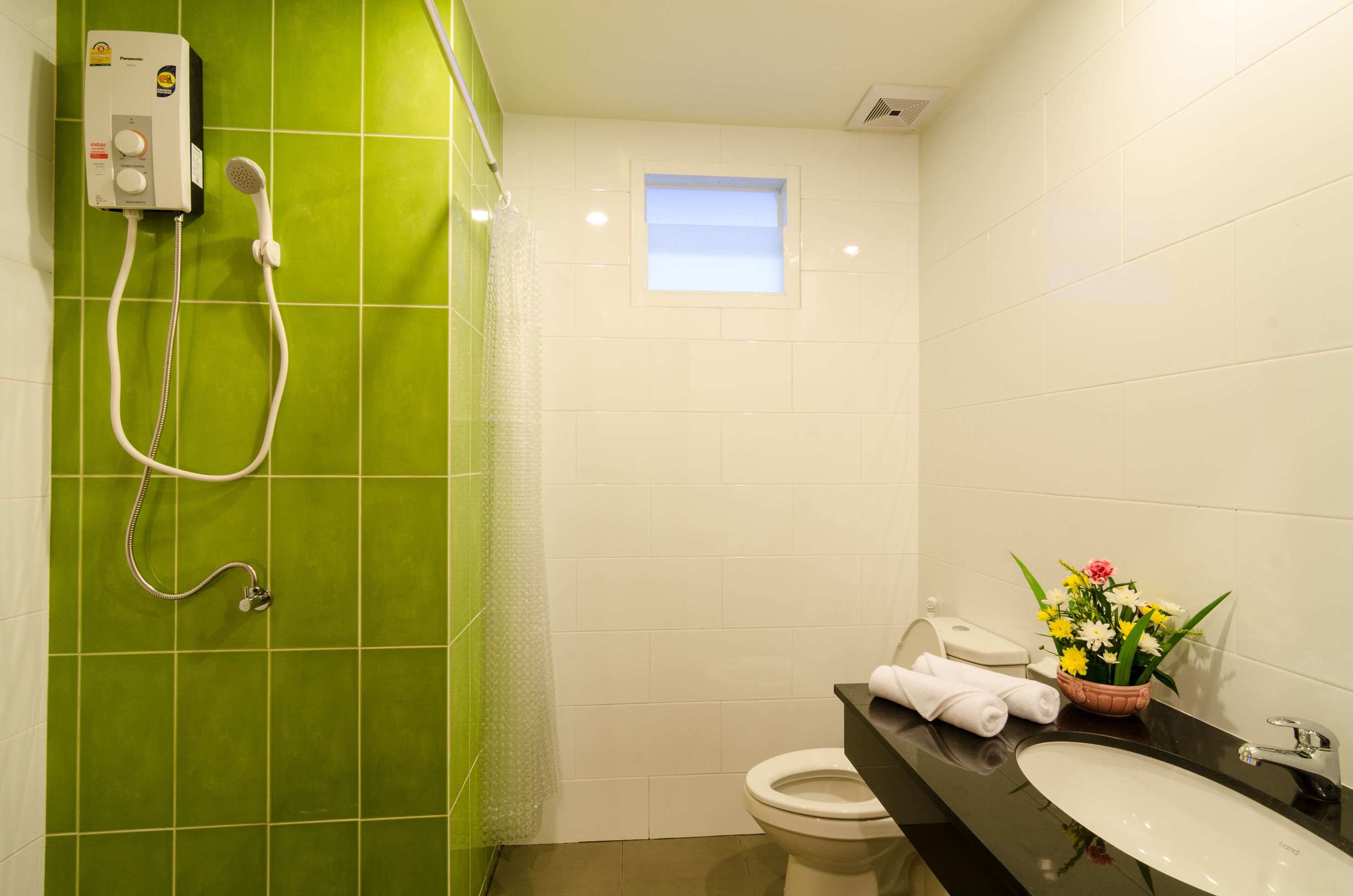 A bathroom with a green tiled wall, a white toilet, a sink with a black countertop, a shower with a handheld showerhead, and a small window.