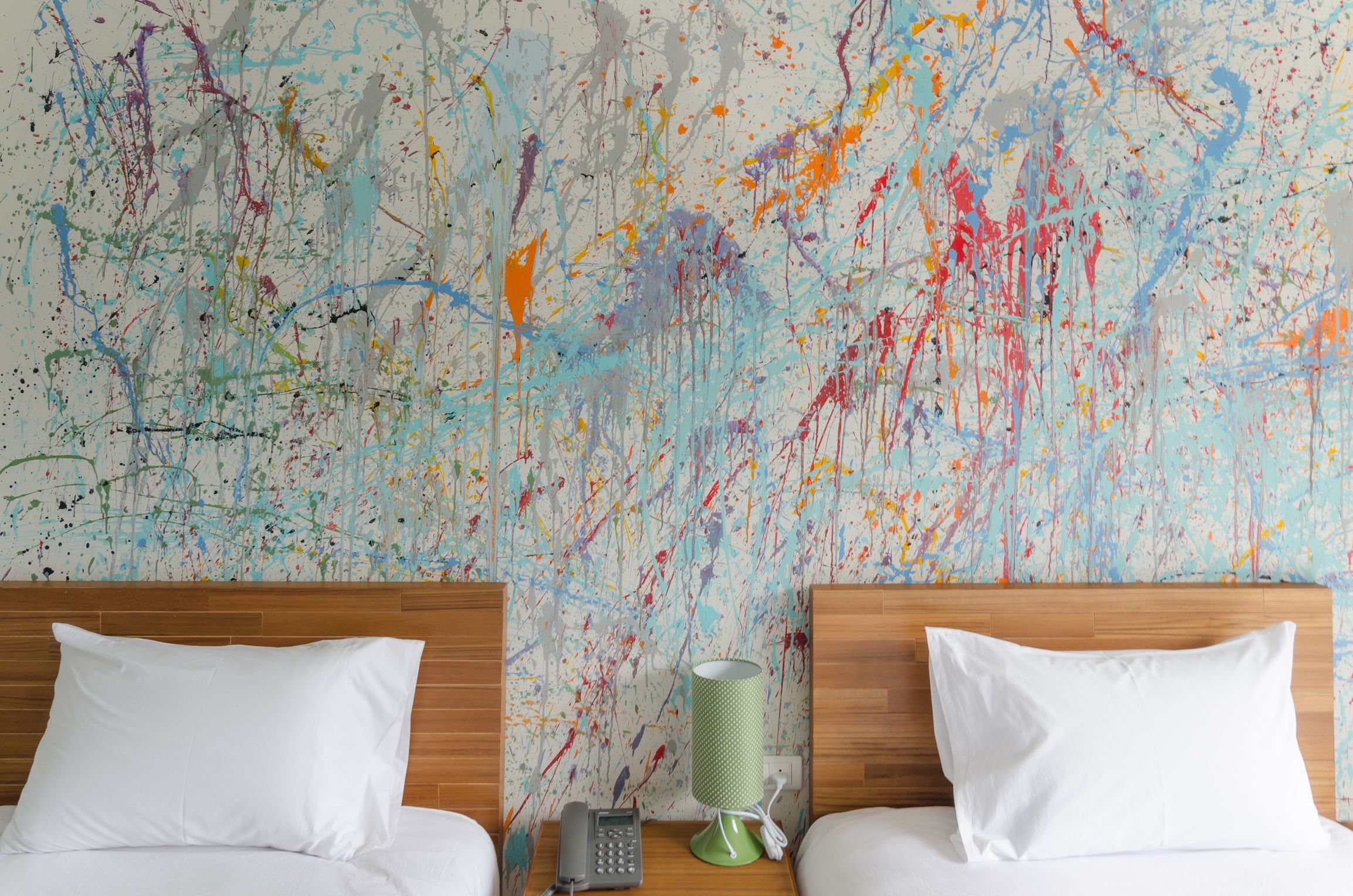Une chambre d’hôtel avec deux lits, des têtes de lit en bois, des oreillers blancs, une lampe verte et un mur orné d’un tableau abstrait aux couleurs vives.