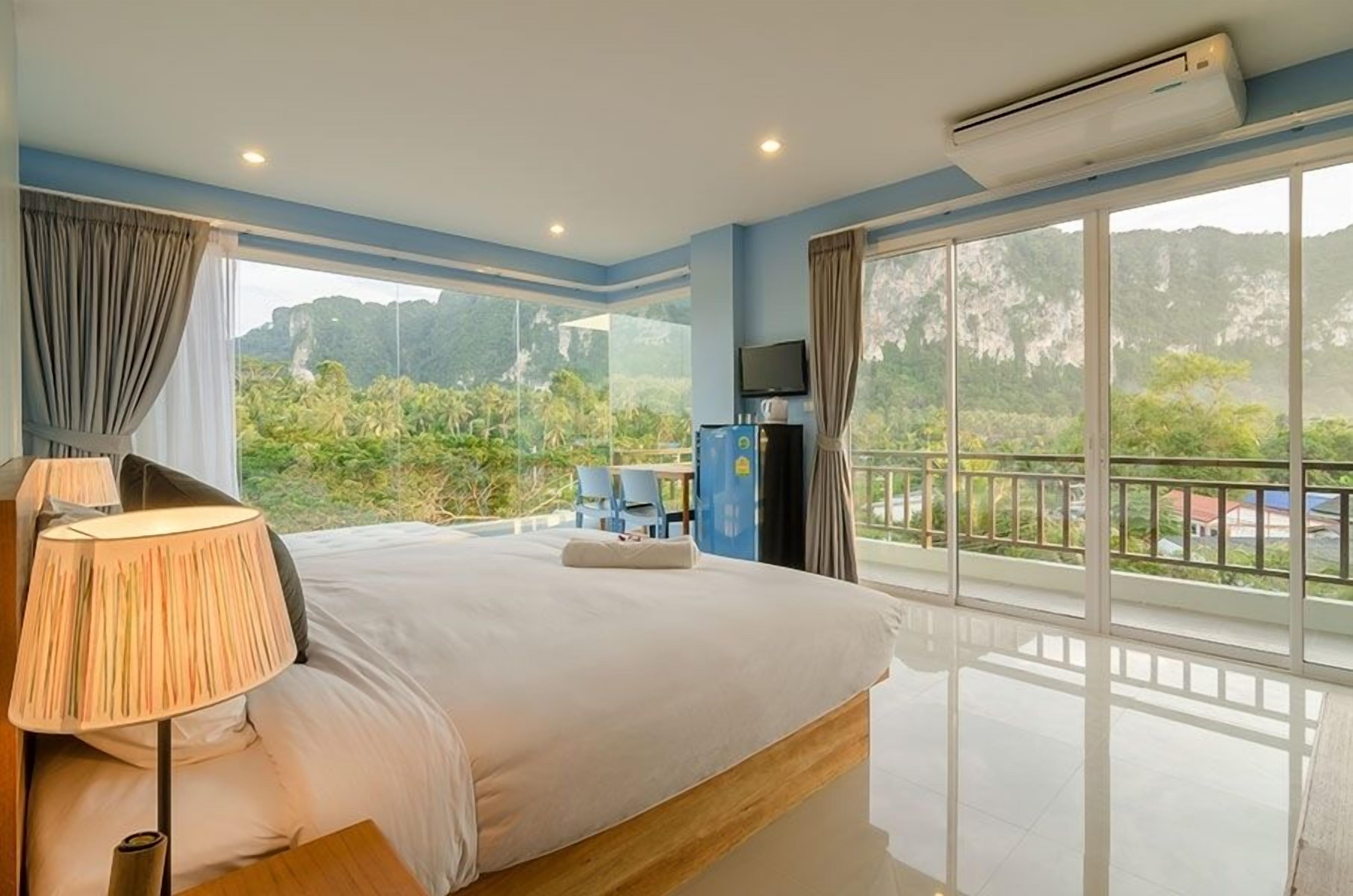 A hotel room with a large bed, a TV, an air conditioner, and a balcony with a view of greenery and mountains.