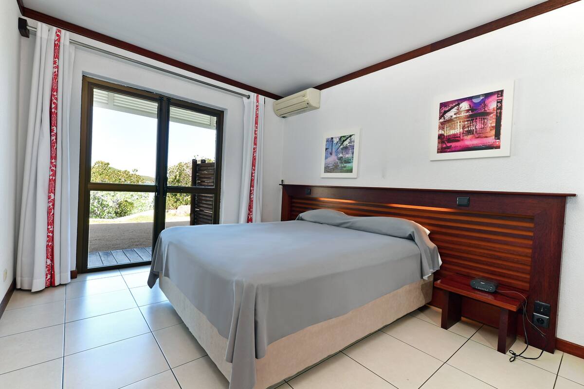 Une chambre comprenant un lit, une table de chevet, un climatiseur fixé au mur et une porte donnant sur un espace extérieur.