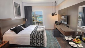 Habitación de hotel con cama, escritorio con televisor, mesa de comedor con fruta y vista a la playa.