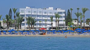 Un hotel con fachada blanca, varios balcones y una zona de playa con sombrillas y tumbonas azules.