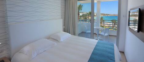 A hotel room with a large bed, a balcony with a view of the beach, and a TV mounted on the wall.
