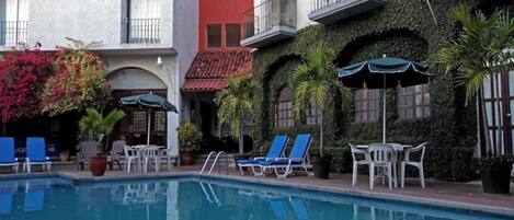 A pool area with lounge chairs and umbrellas, surrounded by buildings with balconies and greenery.