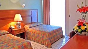 Habitación de hotel con dos camas, una mesita de noche con lámpara, cabecera de madera, ventana con cortinas y un jarrón con flores sobre una mesa.