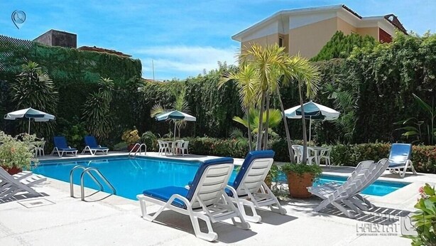 Un área de piscina con sillones de descanso y sombrillas, rodeada de vegetación y un edificio al fondo.