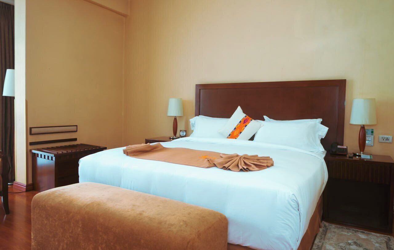 Una habitación de hotel con una cama grande, dos mesitas de noche, un banco y dos lámparas.