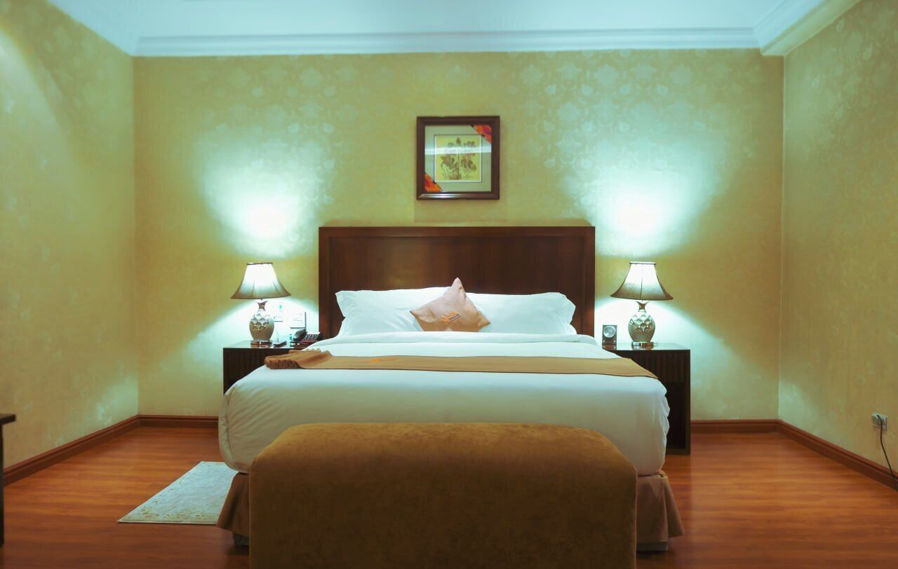A hotel room with a large bed, two bedside lamps, a nightstand with a vase, a framed picture on the wall, and a brown chair.