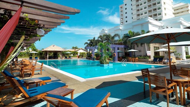 Zona de piscina con sillas de descanso y sombrillas, rodeada por un edificio con balcones.