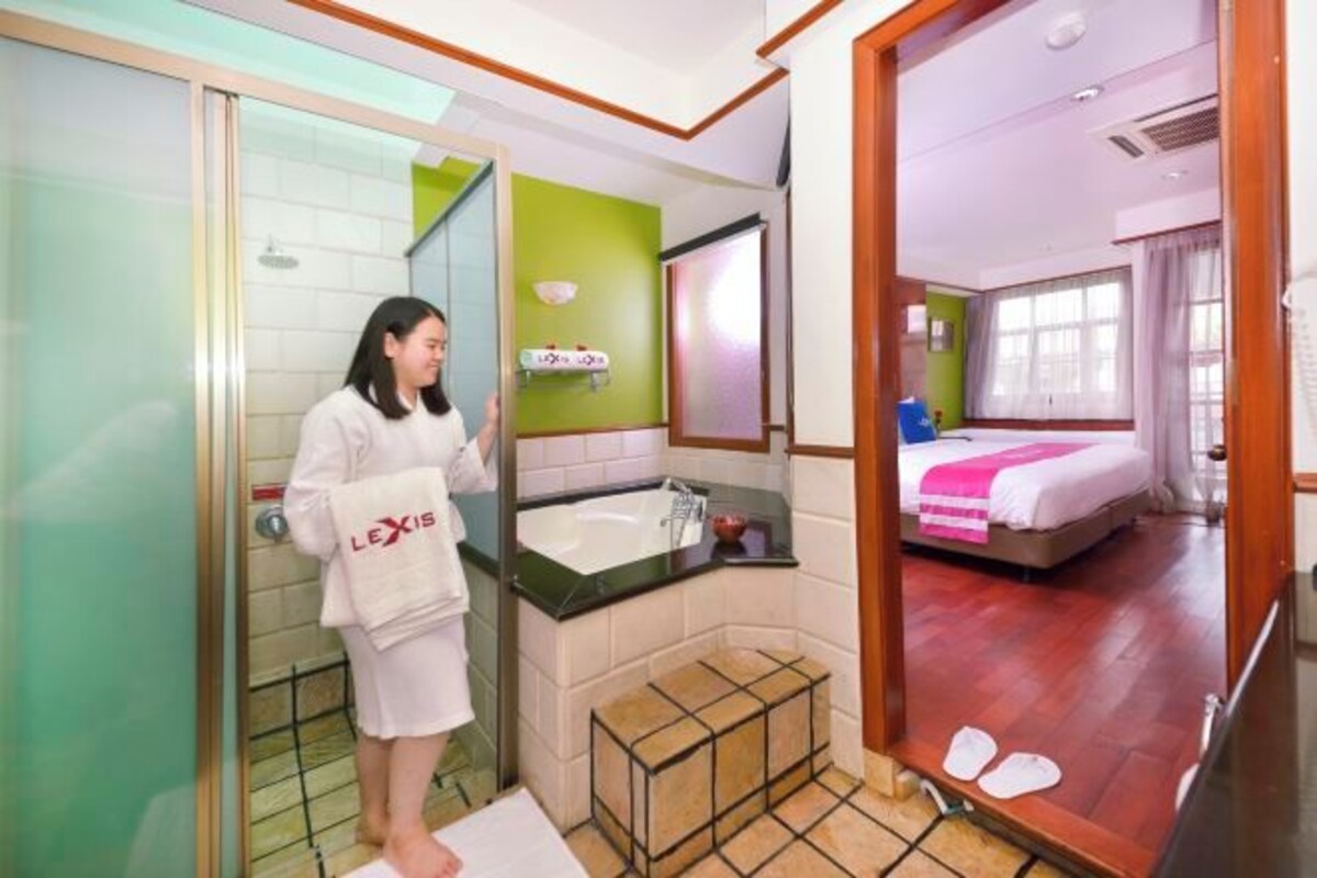 Habitación de hotel con cabina de ducha de vidrio, cama frente a una pared verde y un espejo grande.