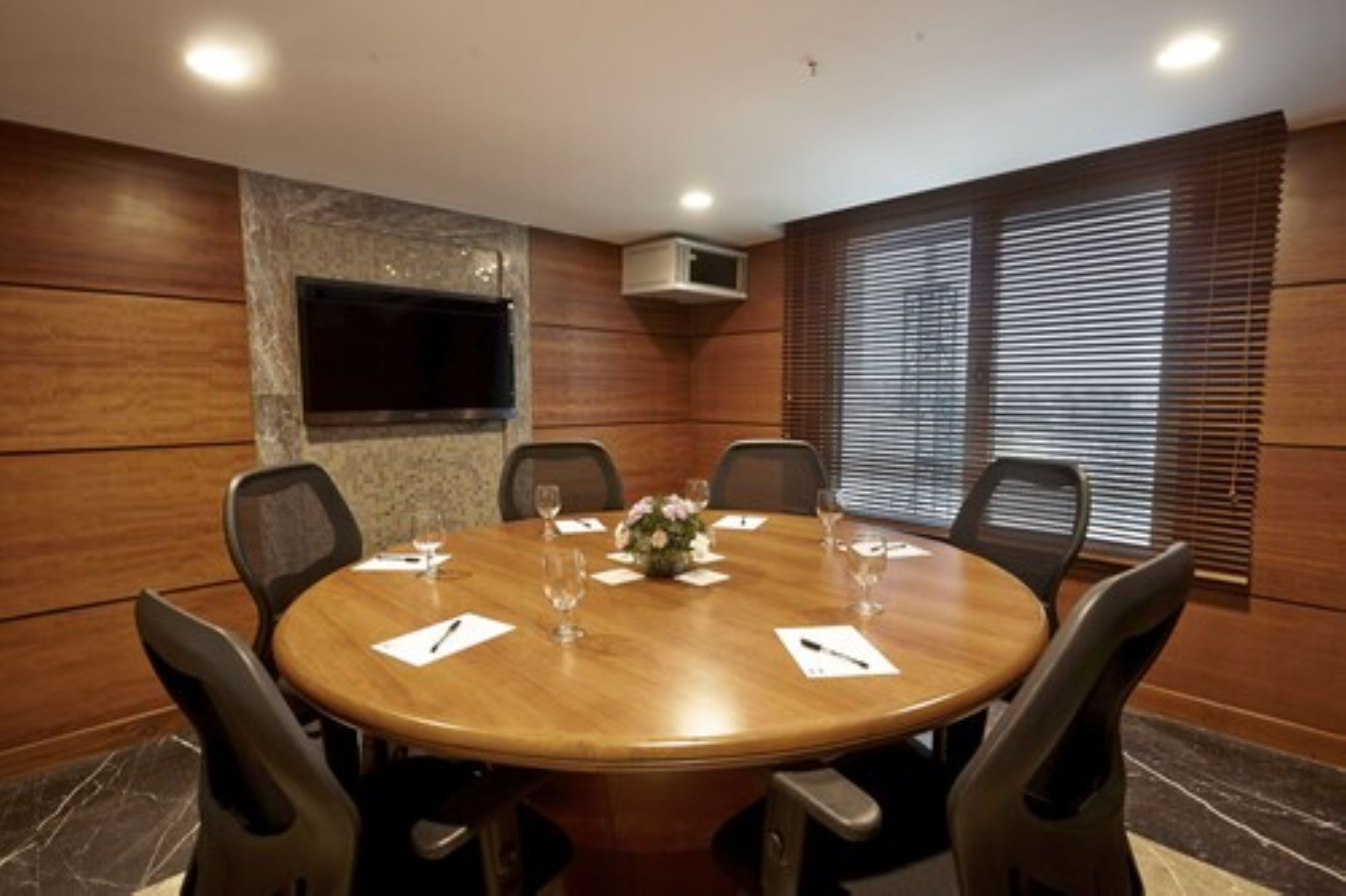 Une salle de conférence avec une table ronde en bois, six chaises, un arrangement floral au centre et un téléviseur fixé au mur.