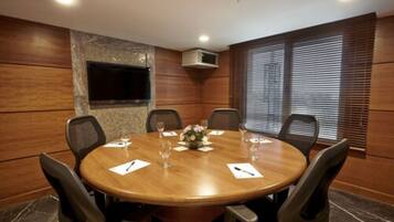 Une salle de conférence avec une table ronde en bois, six chaises, un arrangement floral au centre et un téléviseur fixé au mur.