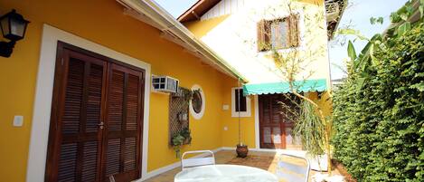 Un edificio amarillo con un patio que cuenta con una mesa redonda y sillas, una planta en maceta y una ventana con una planta de bambú.