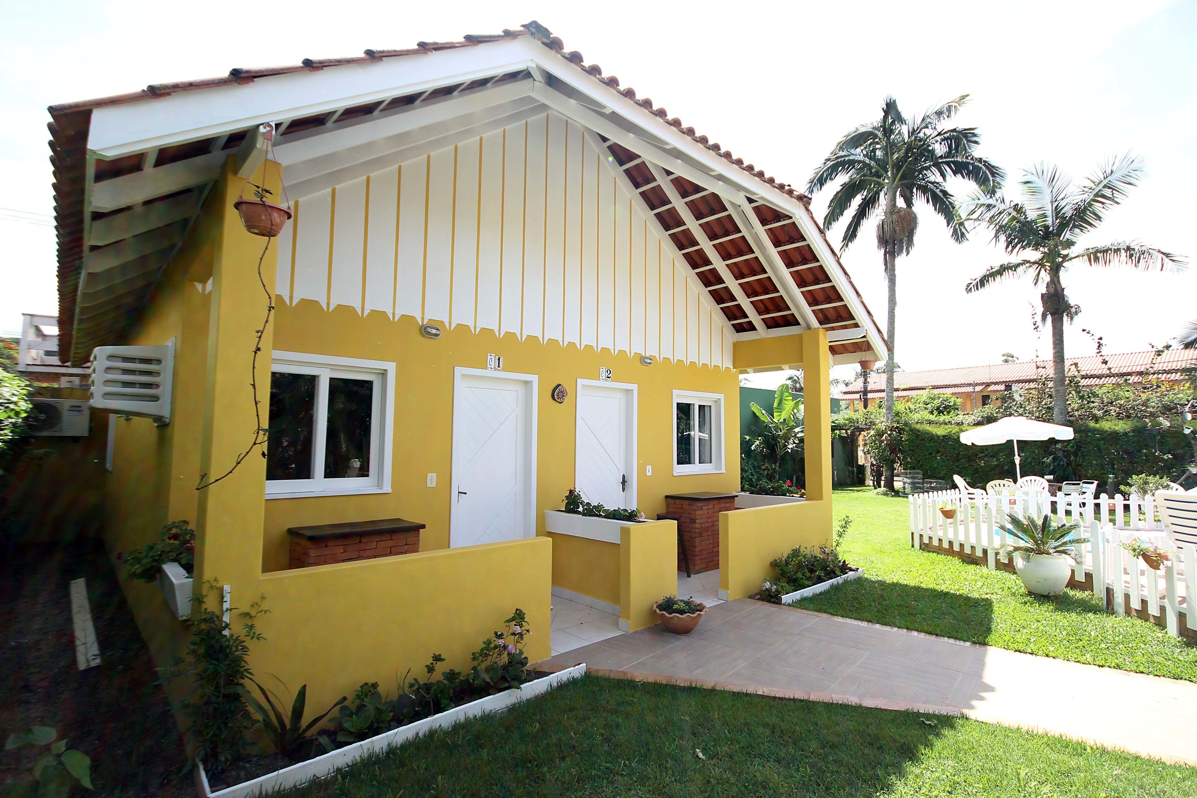 Un edificio amarillo con un porche cubierto, puertas blancas y un pequeño jardín con macetas y una cerca de piquetes blancos.
