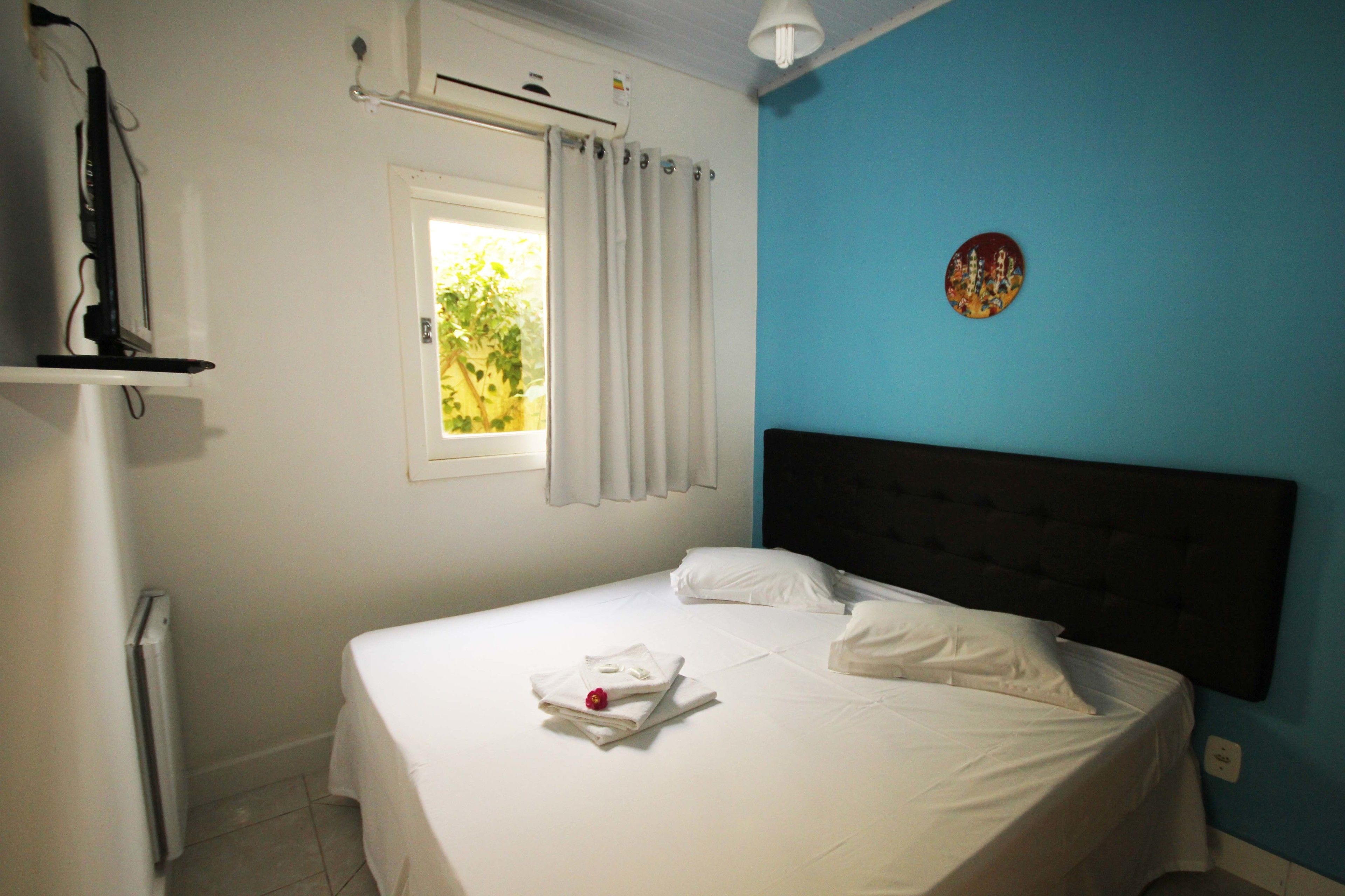 Una cama bien hecha con sábanas blancas y una fruta roja sobre el almohadón, una ventana con cortinas blancas y un aire acondicionado instalado en la pared.