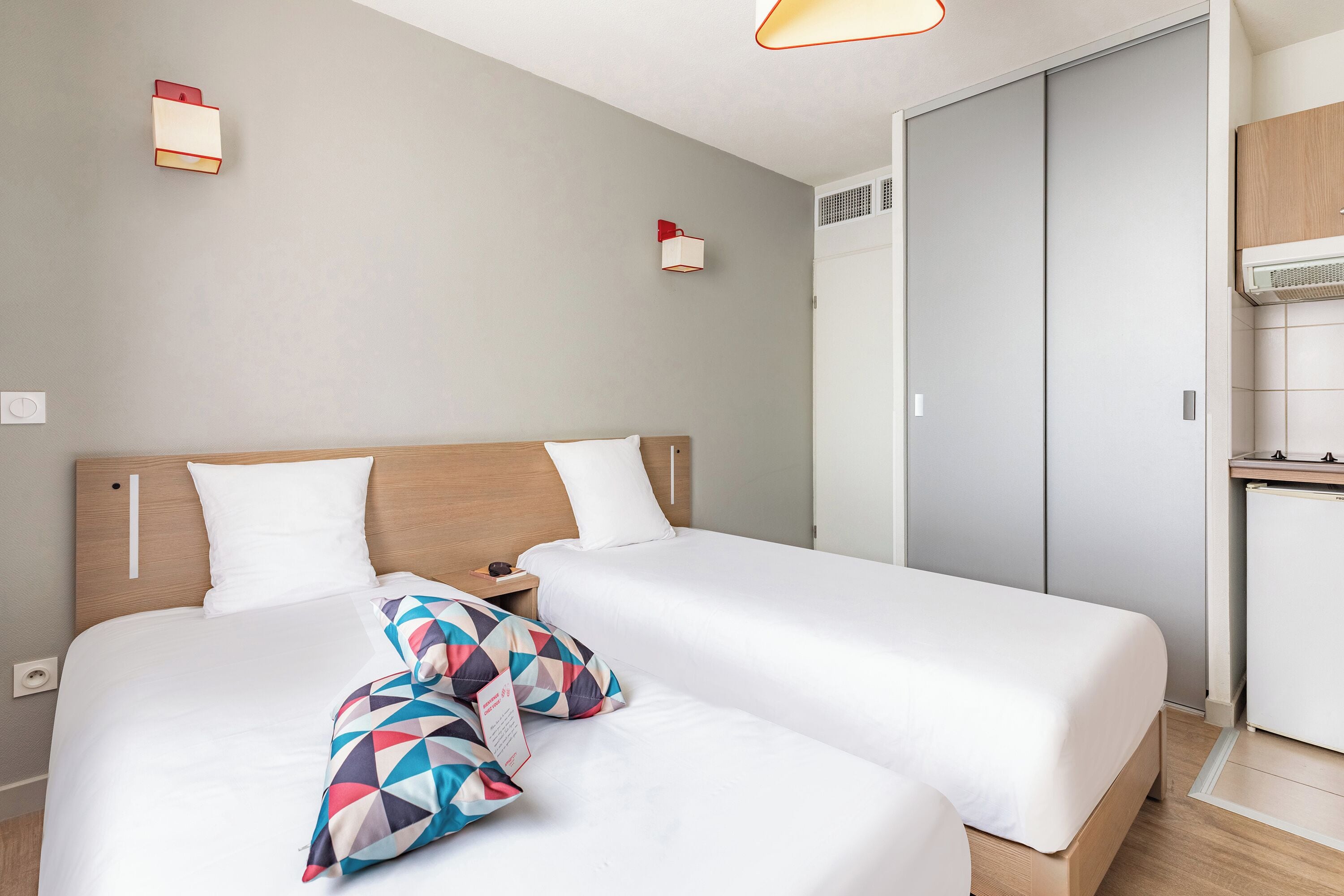 Habitación de hotel con dos camas, cabecera de madera, mesita de noche pequeña y armario empotrado.