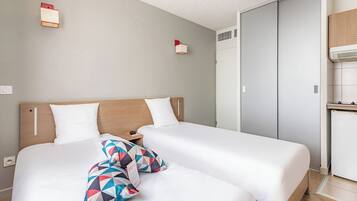 Habitación de hotel con dos camas, cabecera de madera, mesita de noche pequeña y armario empotrado.