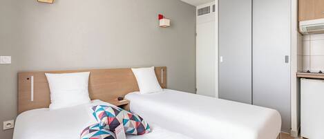 Habitación de hotel con dos camas, cabecera de madera, mesita de noche pequeña y armario empotrado.