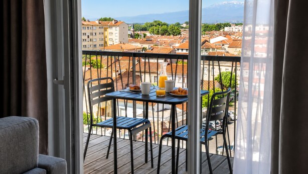 A balcony with a table set for breakfast, chairs, and a view of a cityscape with buildings and mountains.