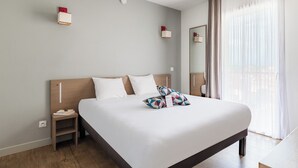 A neatly made bed with white linens and colorful geometric pillows, a bedside table with a book and a phone, and a modern light fixture.