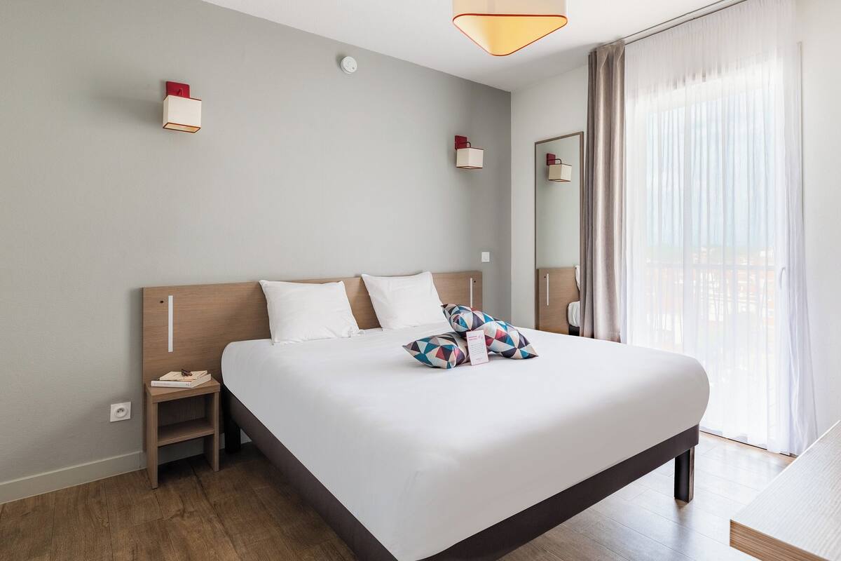A neatly made bed with white linens and colorful geometric pillows, a bedside table with a book and a phone, and a modern light fixture.